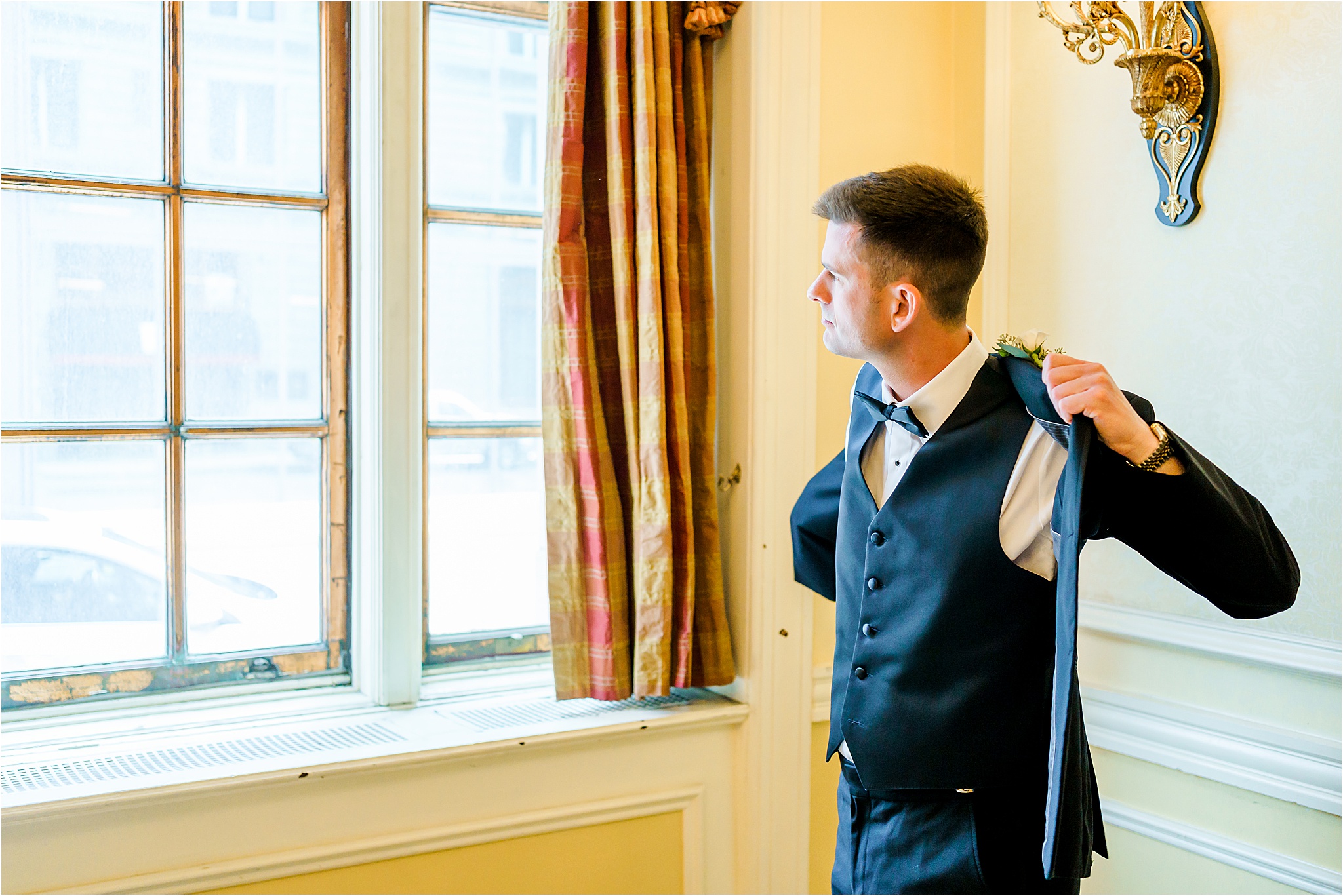 A groom putting on his suit jacket while looking out the window.