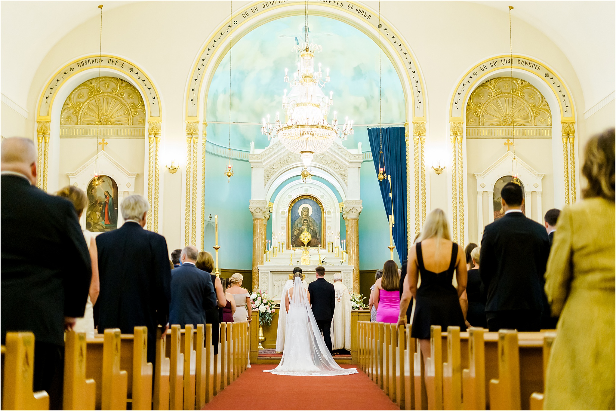 The wedding couple stands at the front of the altar at the church. You can see the backs of guests and the artwork and architecture of the church.