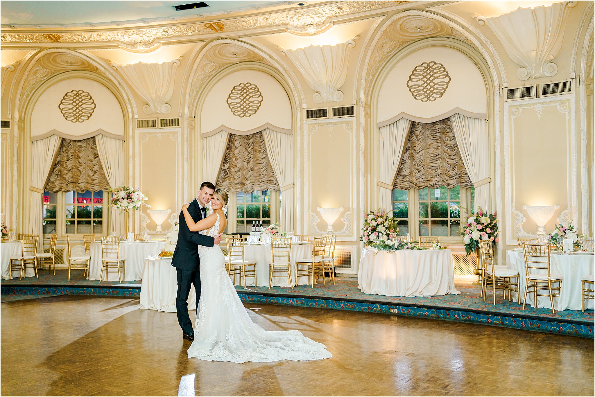 The couple stands facing and holding each other while smiling at the camera. They are standing in a grand ballroom at the Fairmont Copley with tall ceilings and detailed architecture. 