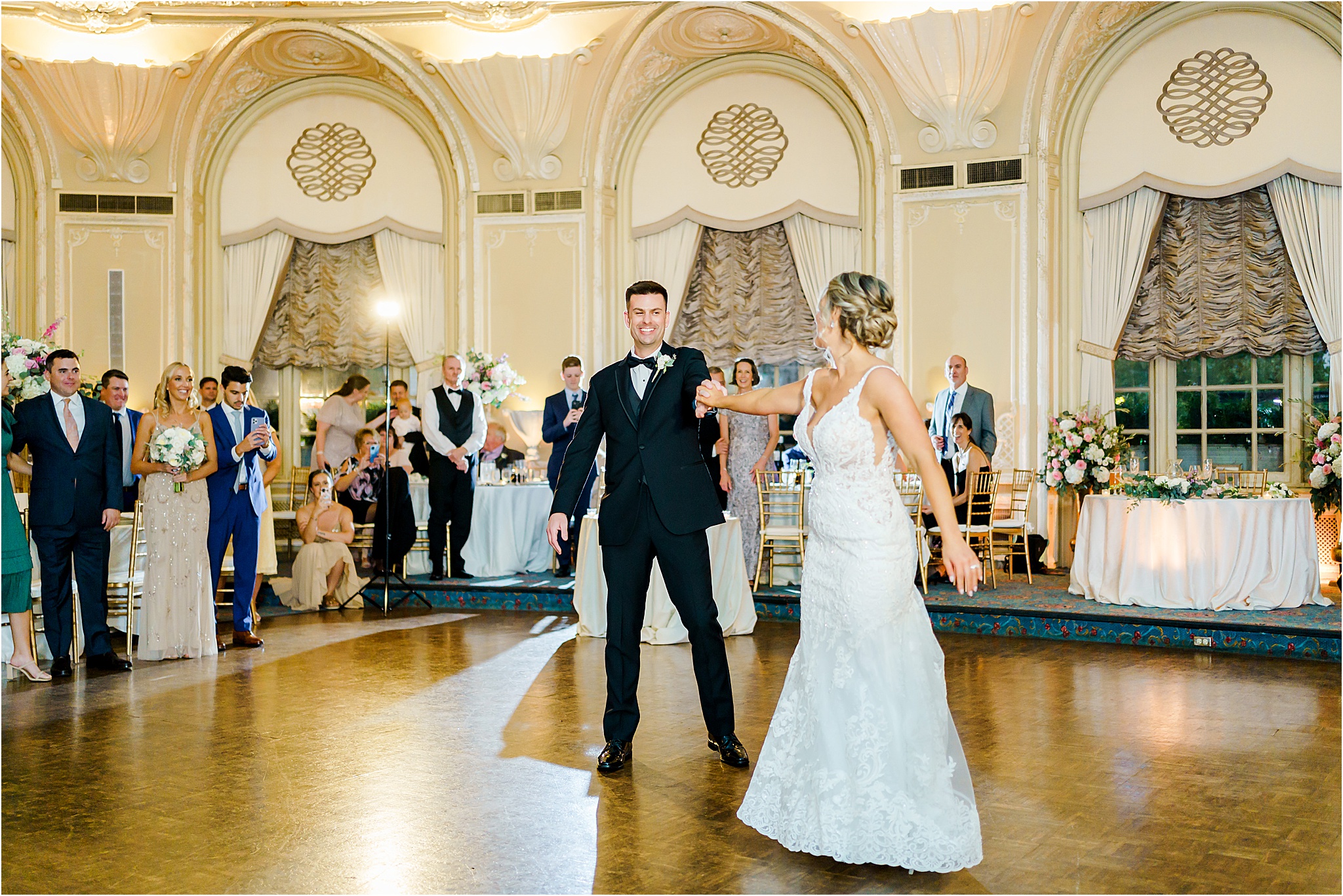 A photo of the couple's first dance. The couple is in the middle of the dance floor with the groom smiling at the bride as he holds her hand after spinning her away from him. The wedding guests can be seen smiling and watching in the back.