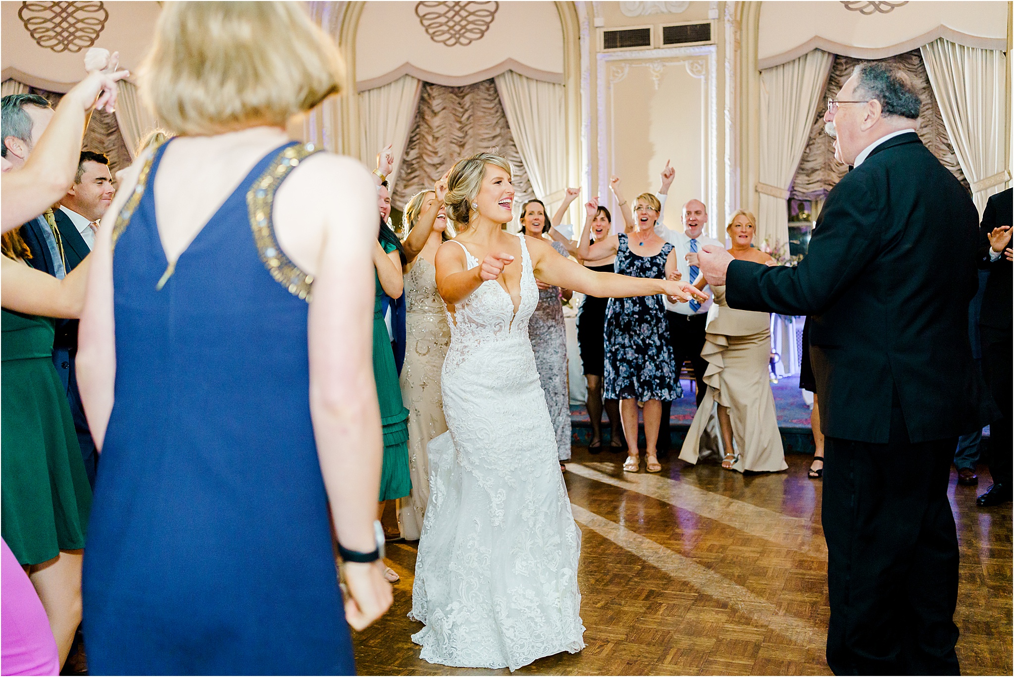 The bride dancing with her arms outstretched and singing to her father while guests crowd around her.