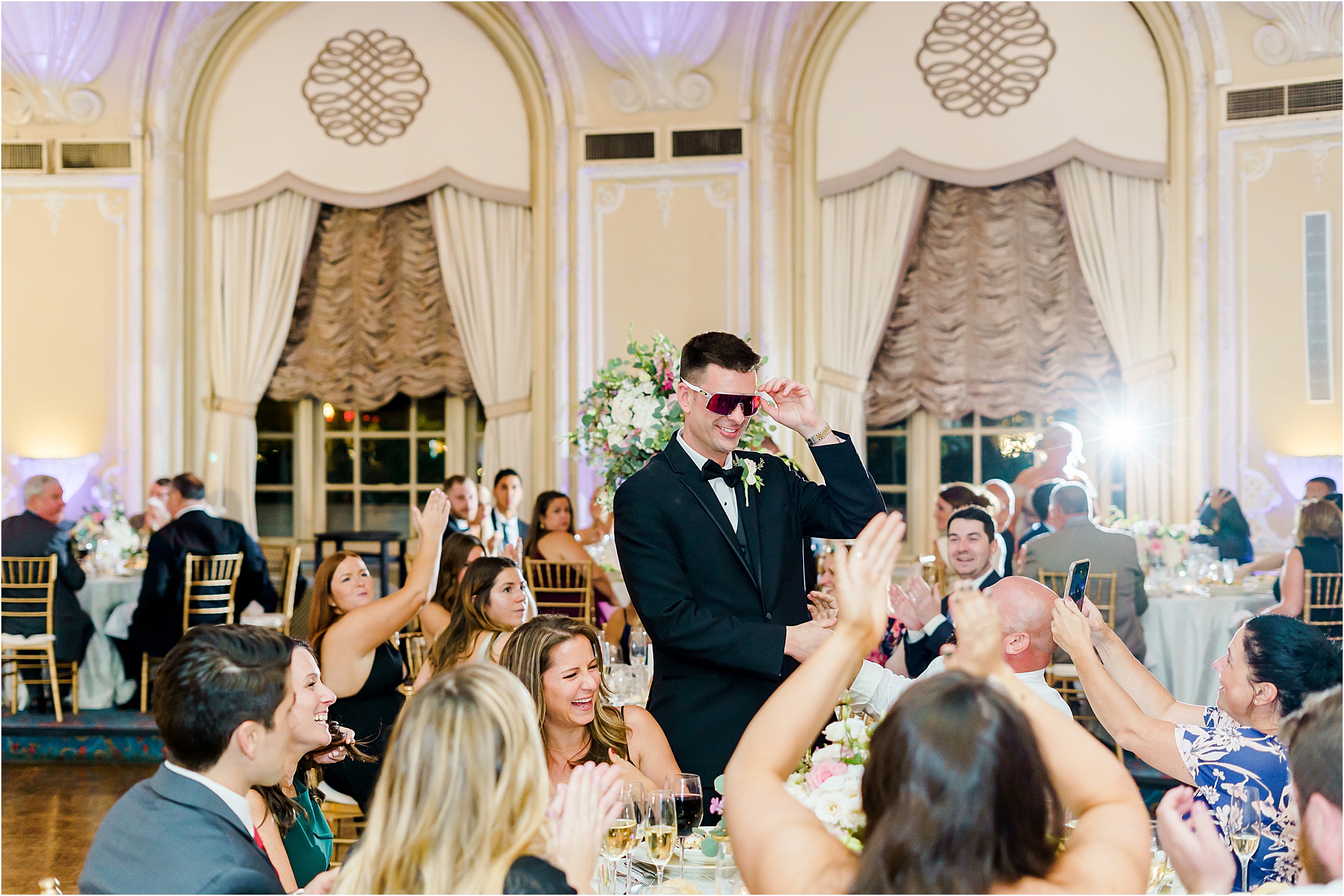 The groom walking through the crowd of guests seated and cheering at their tables. He is wearing dark sunglasses and smiling.