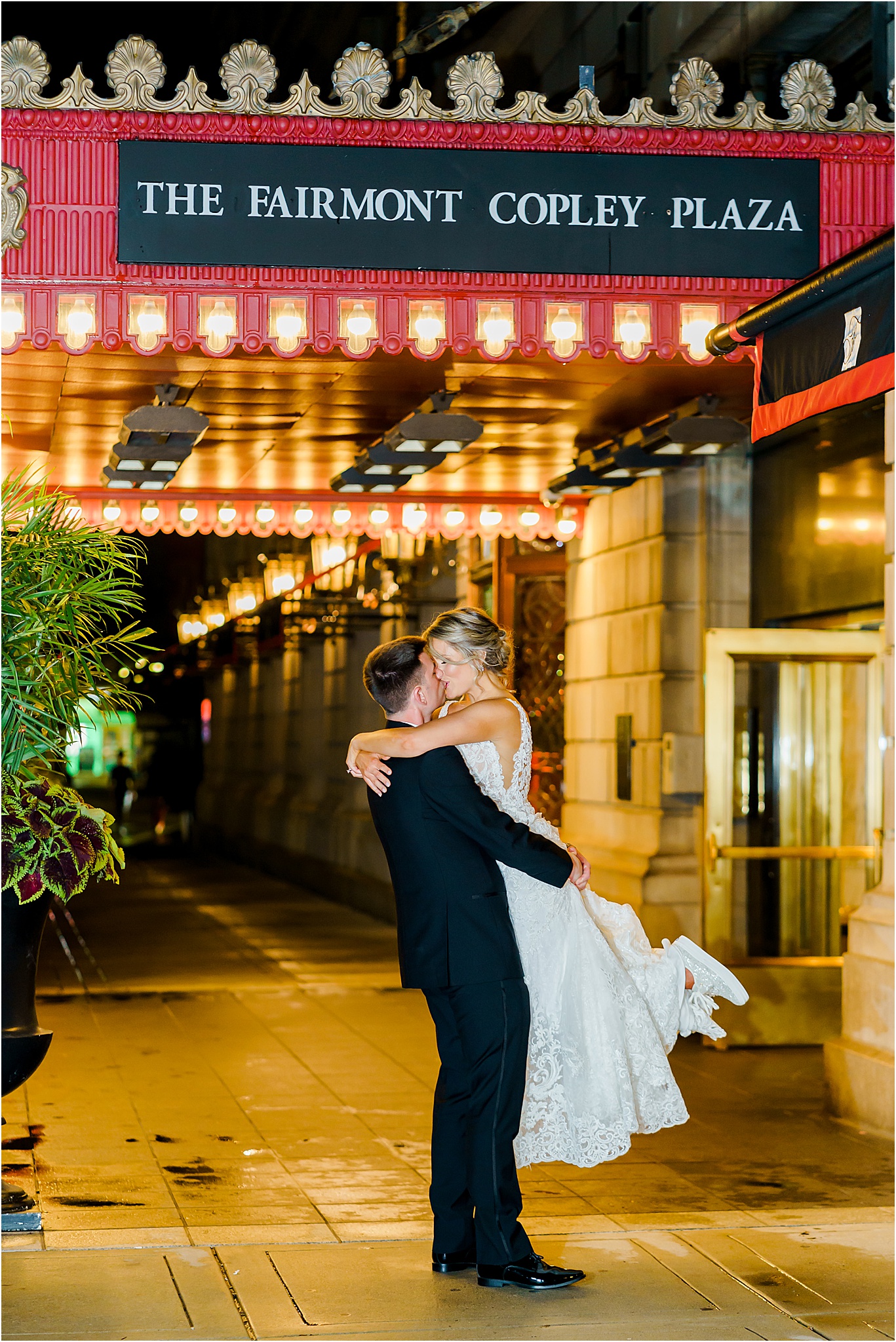 The groom kissing and lifting the bride off the ground while posing for a nighttime portrait under the Fairmont Copley Boston sign and overhang.